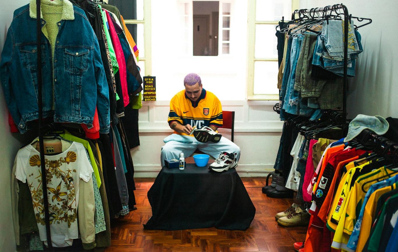 Man working on shoe repair in a vintage clothing store surrounded by colorful attire.
